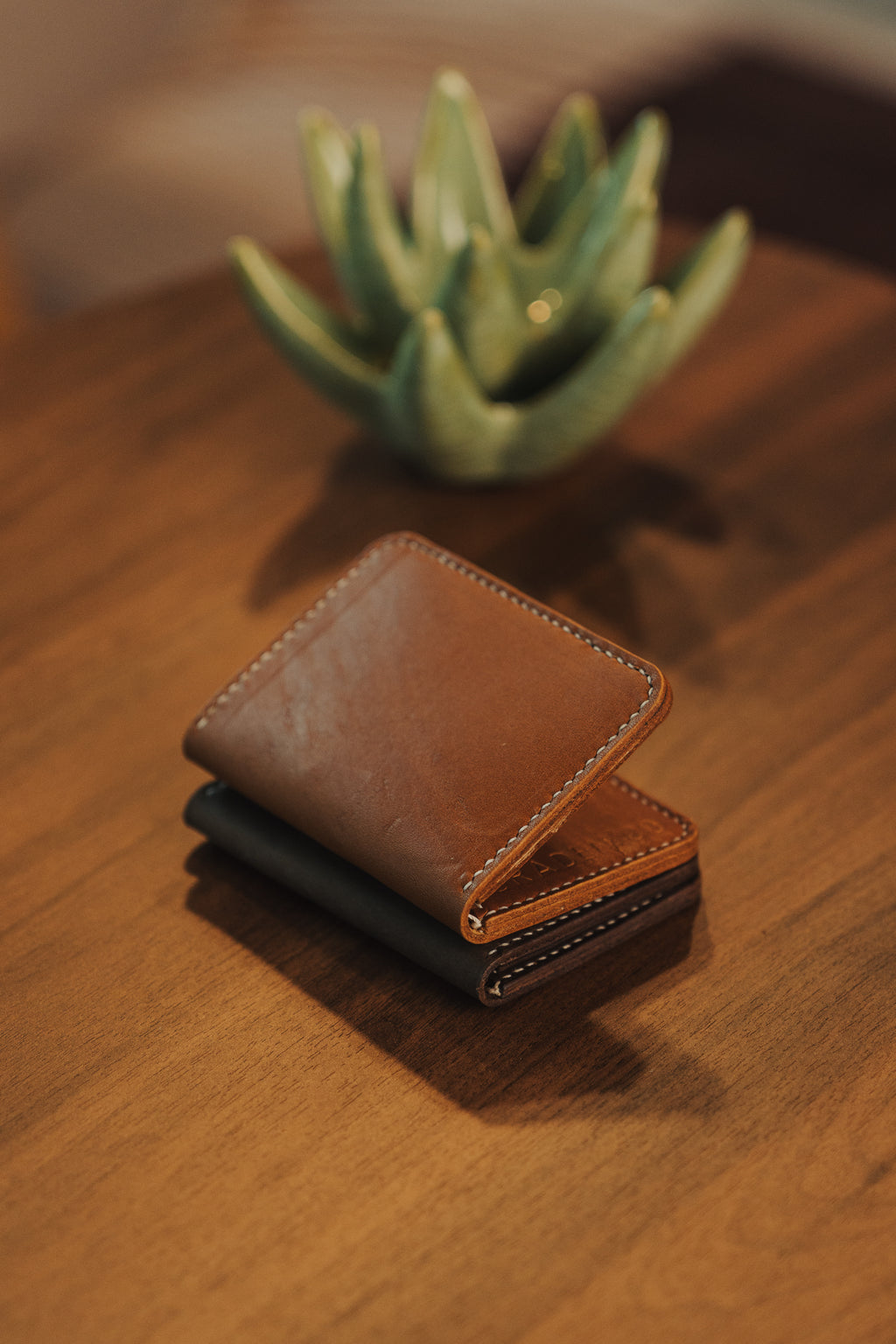 Two leather wallets stacked on a wooden surface with a plant in the background