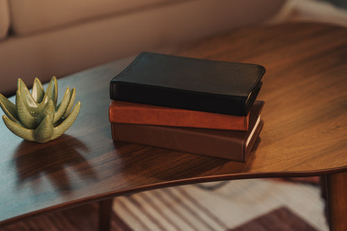 Three books stacked on a wooden table with a small green plant beside them.