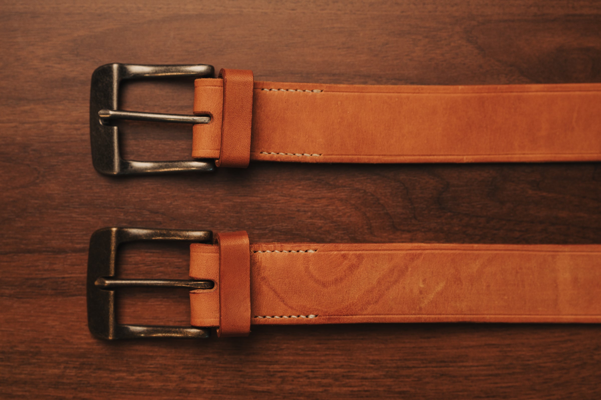 Two brown leather belts with one brass and one silver buckle on a wooden surface